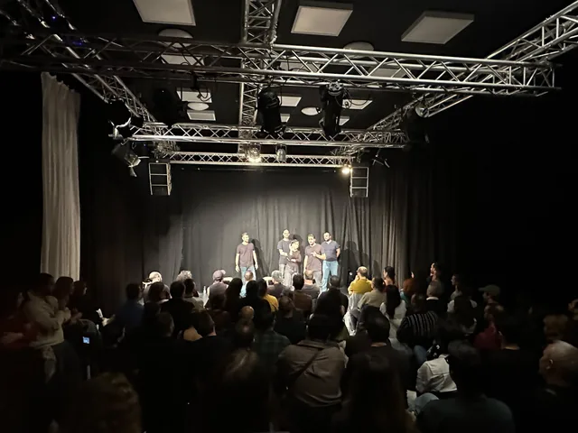 Publikum in einem vollen Saal und fünf Darstellende gemeinsam auf der Bühne