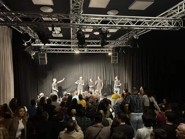 Blick von hinter den Publikumsreihen auf die beleuchtete Bühne und das Ensemble